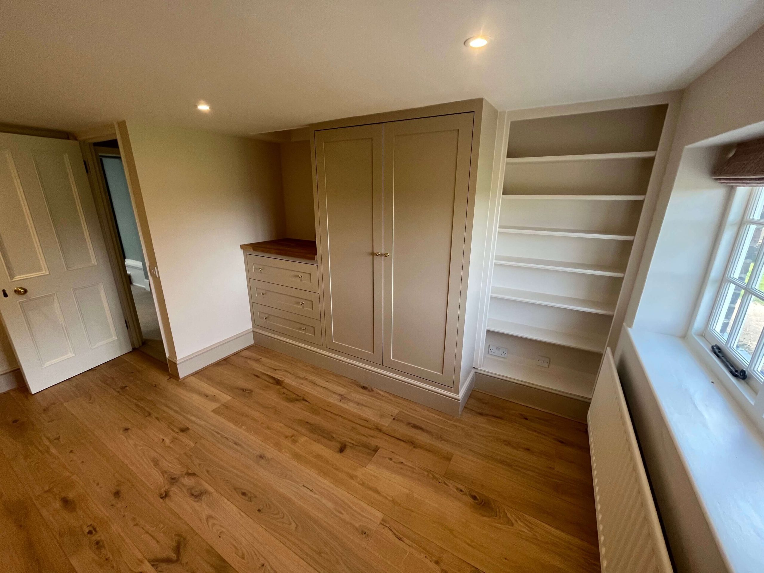 Fitted bedroom wardrobe in painted greige with oak-topped drawer unit and alcove shelving, on an oak floor