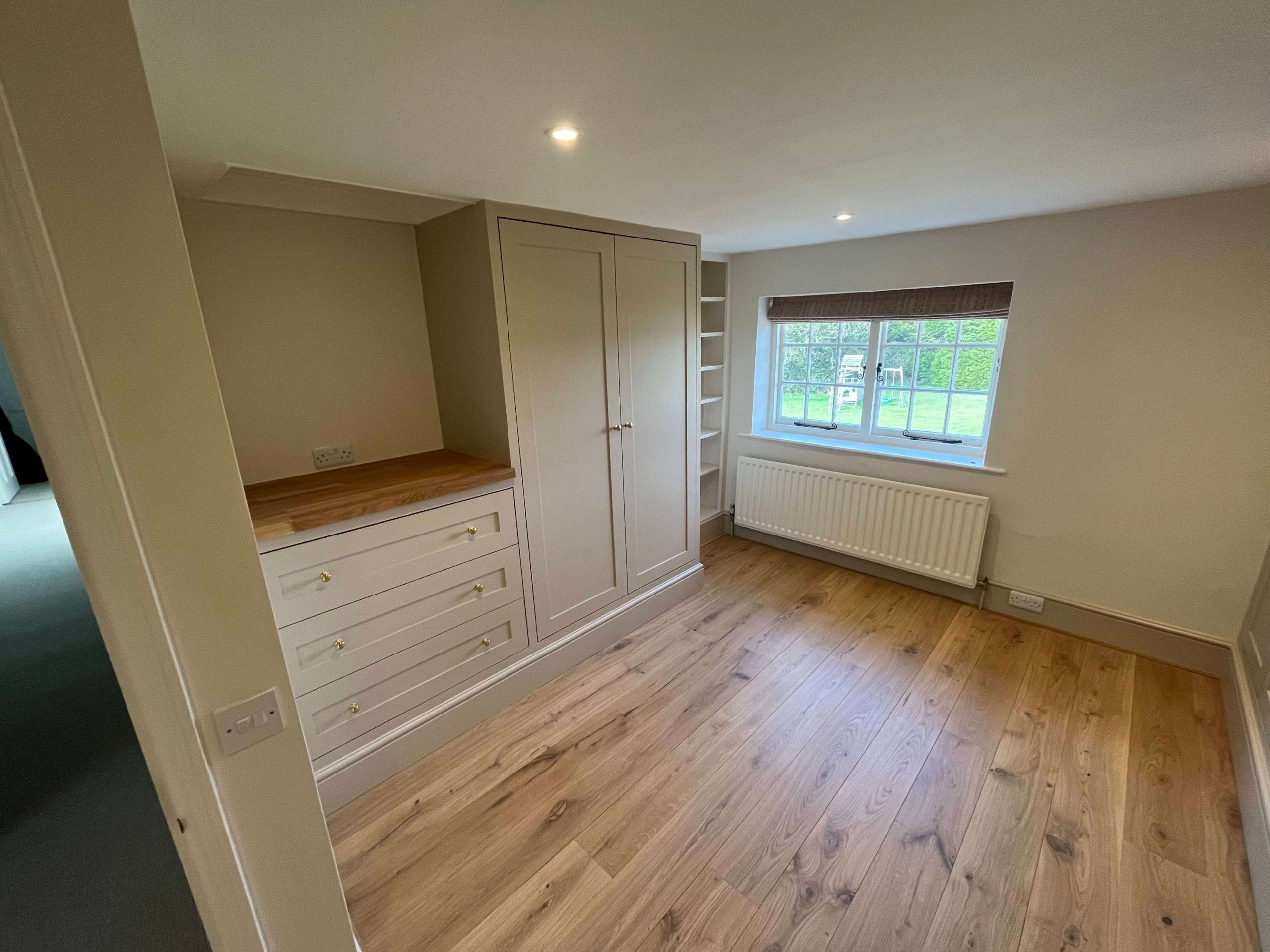 Built-in bedroom storage with painted wardrobe, oak-topped drawers and open shelving beside a window